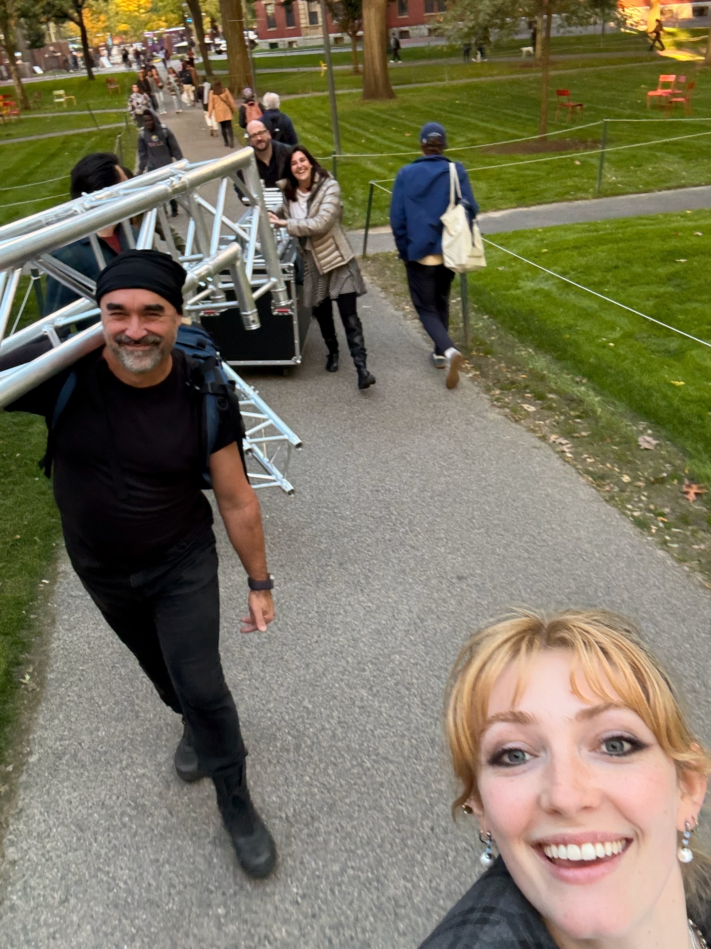 a group of Learning Lab staff and fellows smiling and carrying truss and large media equipment walking through a grassy university campus yard