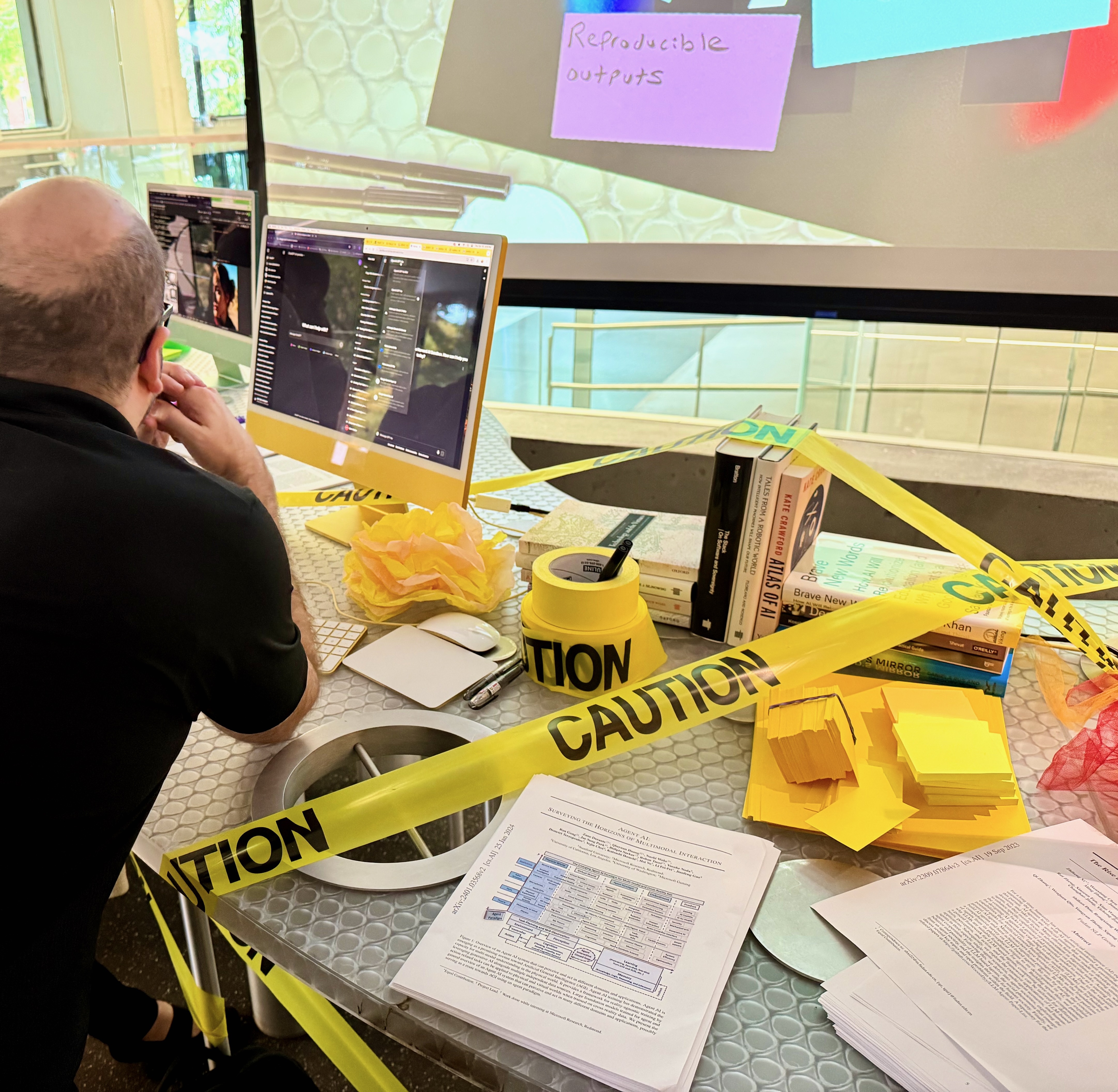 Learning Lab staff looking at the yellow computer screen with different AI tools open. The tabletop he is leaning on has caution tape, articles, and books on it.
