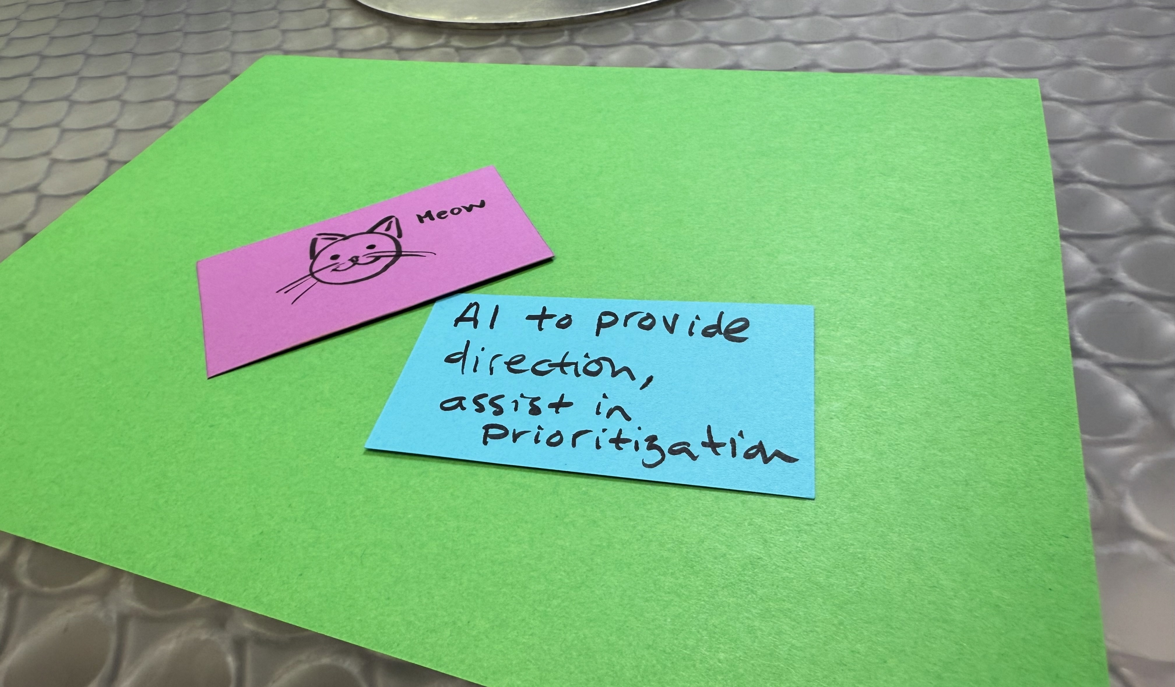 two cards on a green piece of paper, one card is purple and reads "meow" with a picture of a cat, and the other card is blue and reads "AI to provide direction, assist in prioritization"