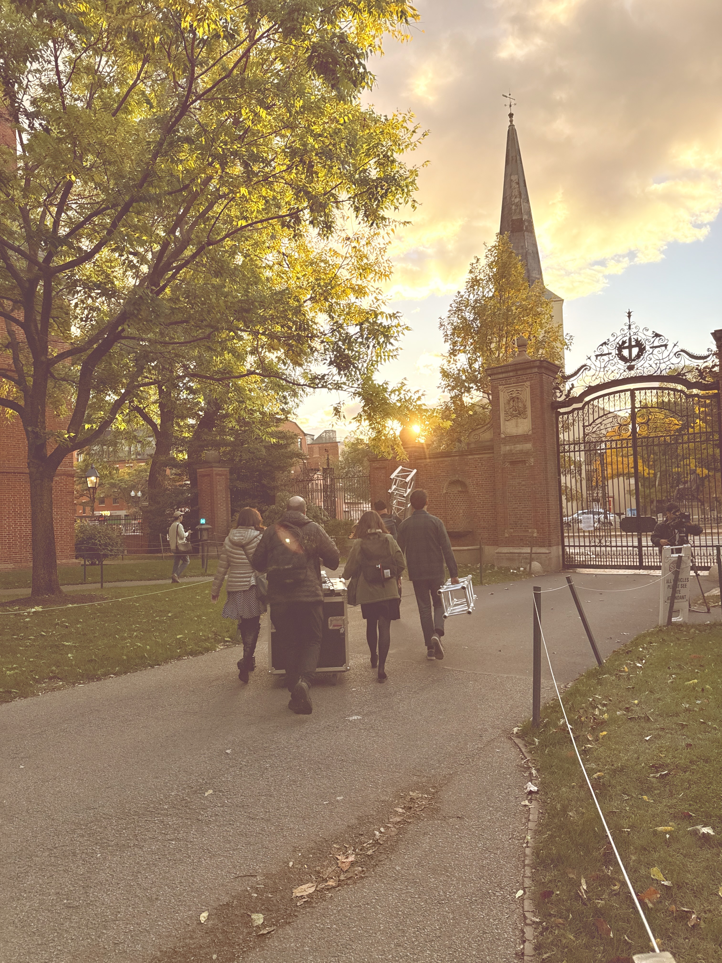 a group of Learning Lab staff and fellows carrying truss and large media equipment, walking into a sunset with a church on the right-hand side