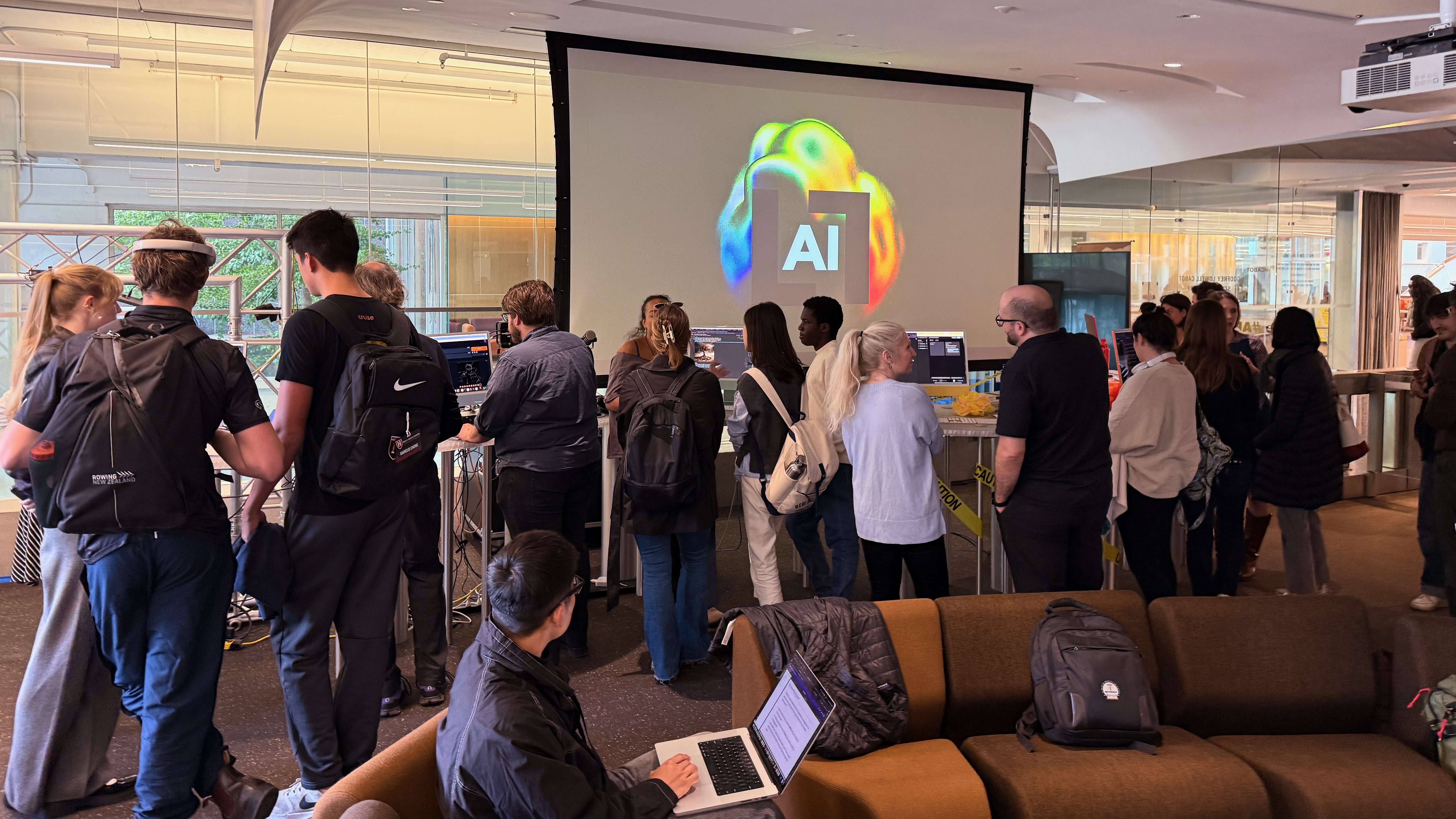 a large group of people speaking in smaller groups in front of the rainbow of iMacs and a screen that says "LL" and "AI"